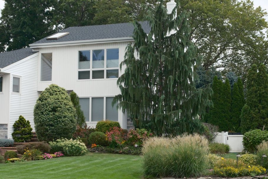 White two-story house with large windows, landscaped flower beds, and tall trees in the yard