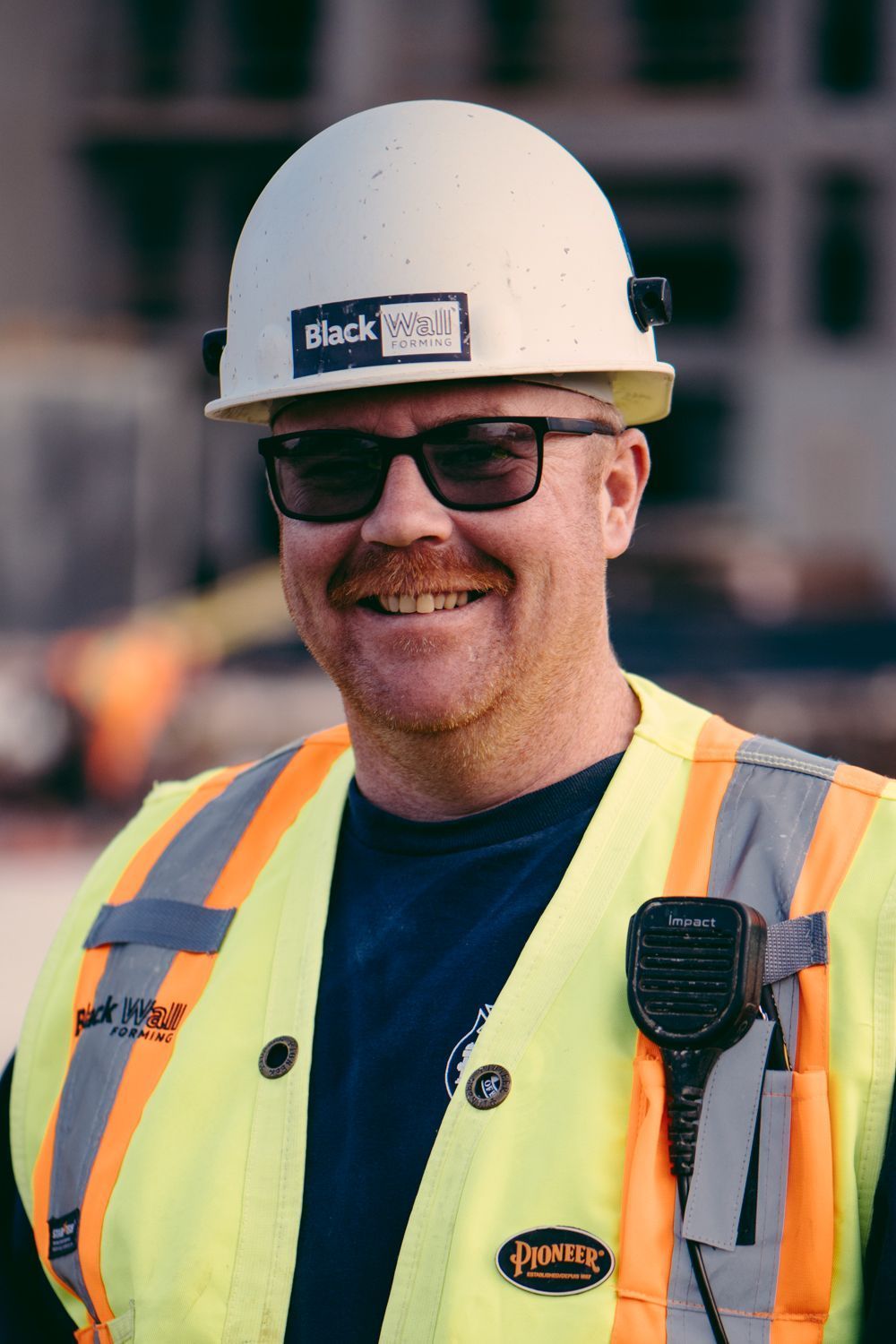 Construction worker in white hard hat, glasses, and orange safety vest, smiling at a job site