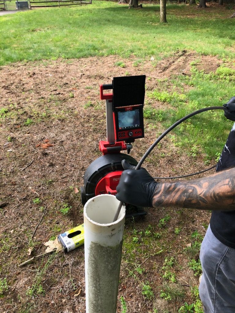 Worker using a pipe inspection camera beside a white PVC pipe in a muddy yard