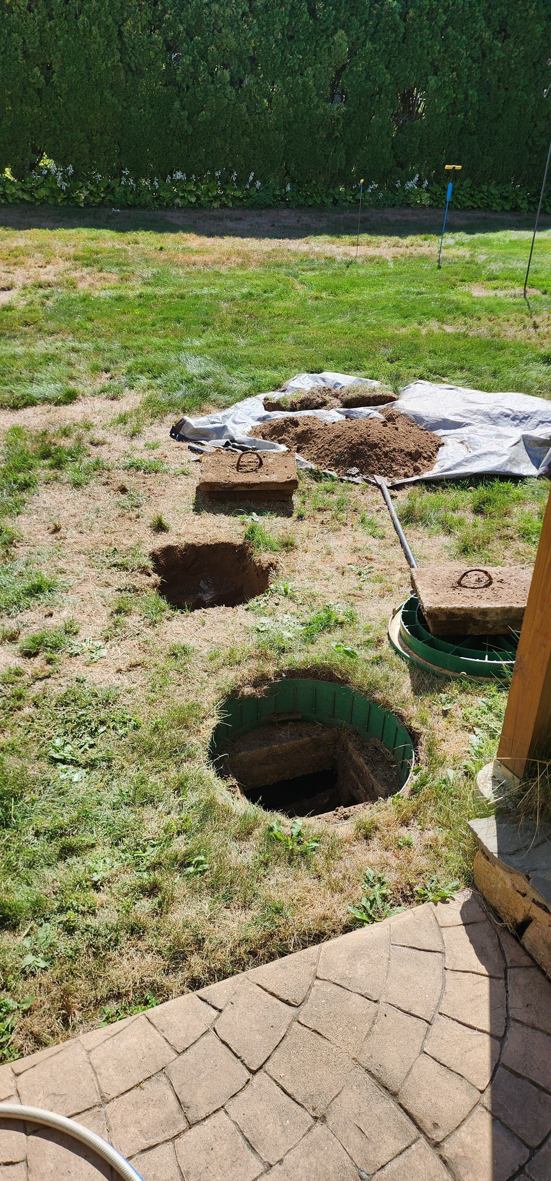 Backyard with two open dirt pits, a tarp-covered area, and a paved patio edge