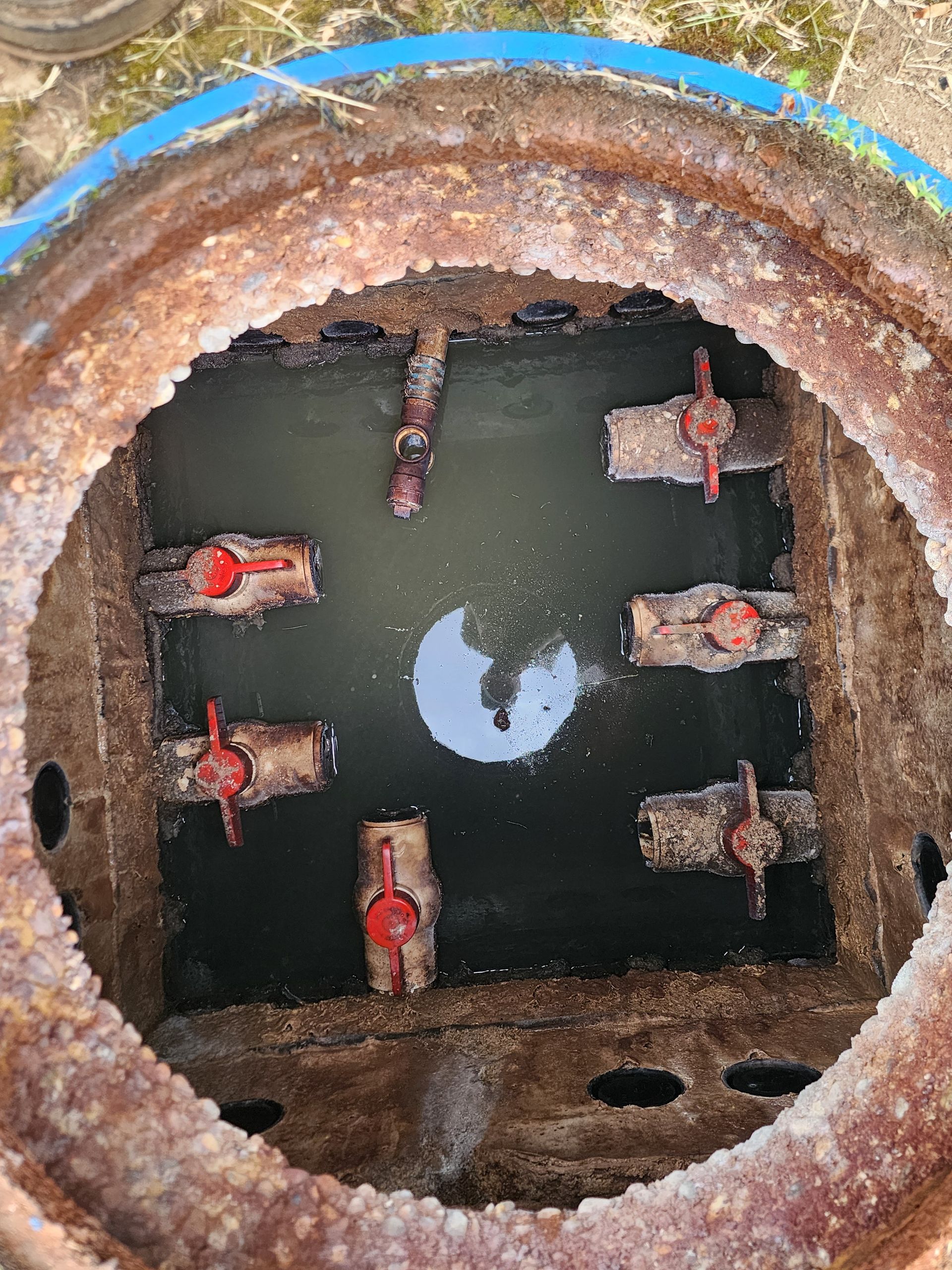 Rusty circular manhole with open water and several red valves inside concrete shaft