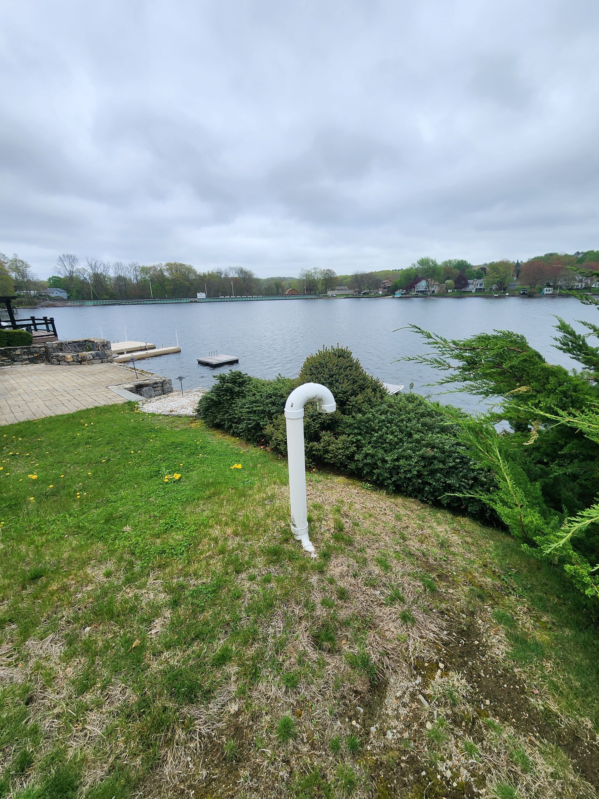 Grassy lakefront with a white vent pipe, shrubs, rocks, and cloudy sky over calm water