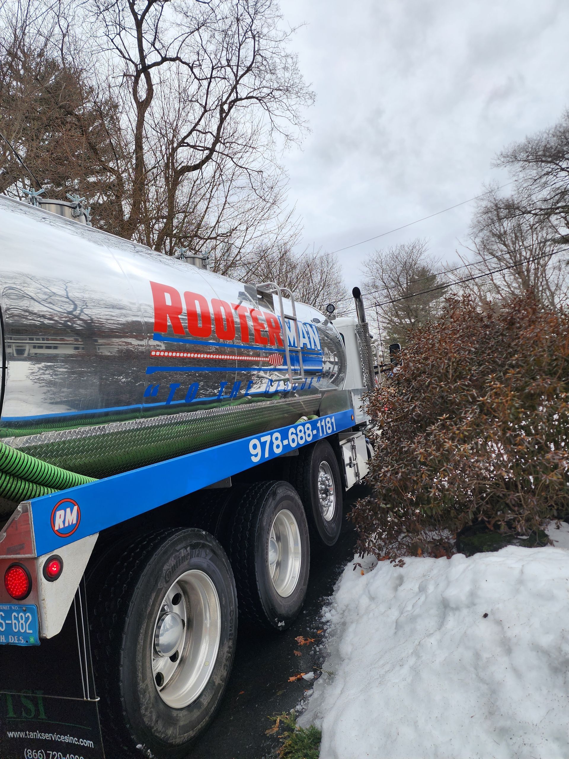 Blue and silver tanker truck parked beside snowbanks on a snowy roadside with trees in the background