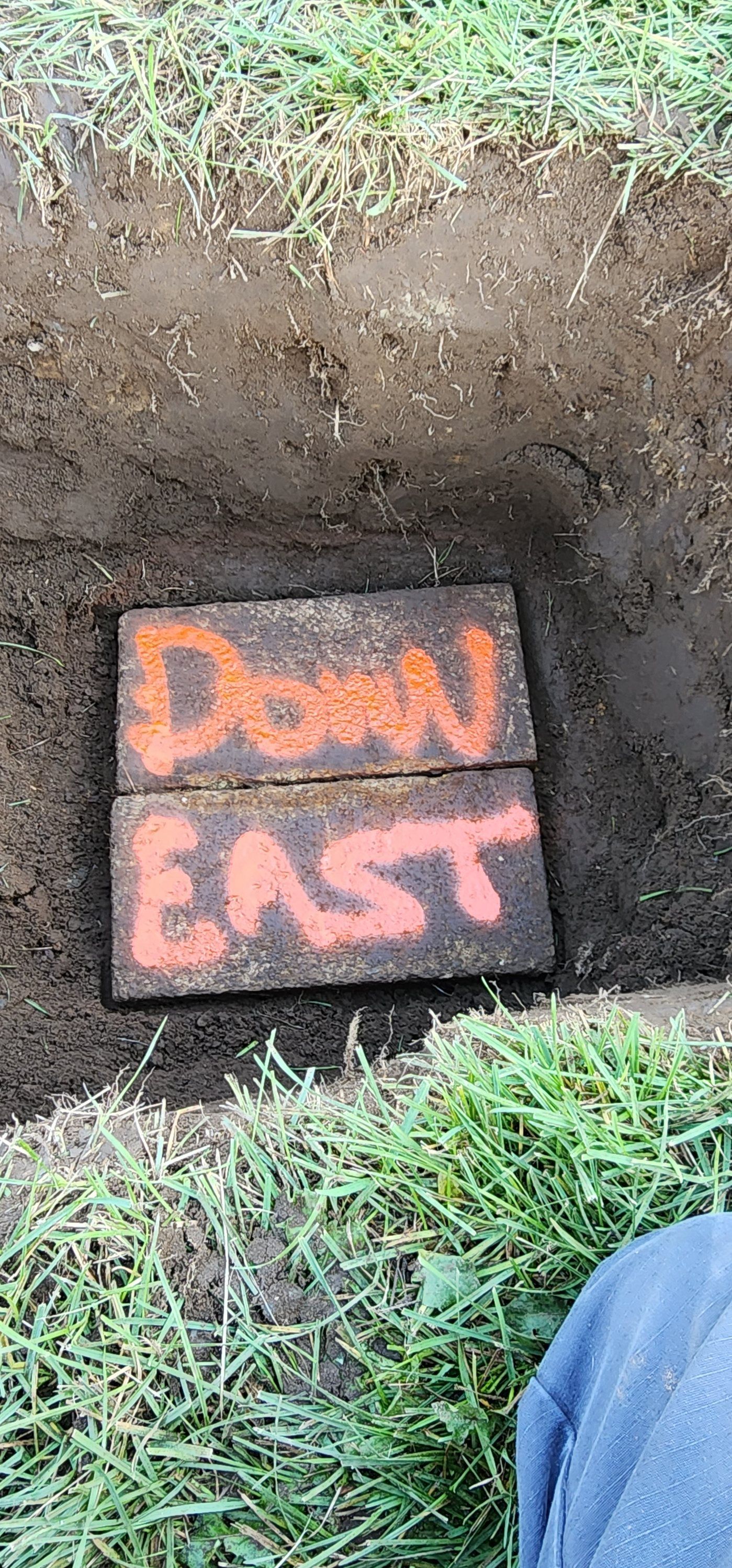 Orange “DOWN EAST” sign on the ground beside a dug-out dirt hole and grass