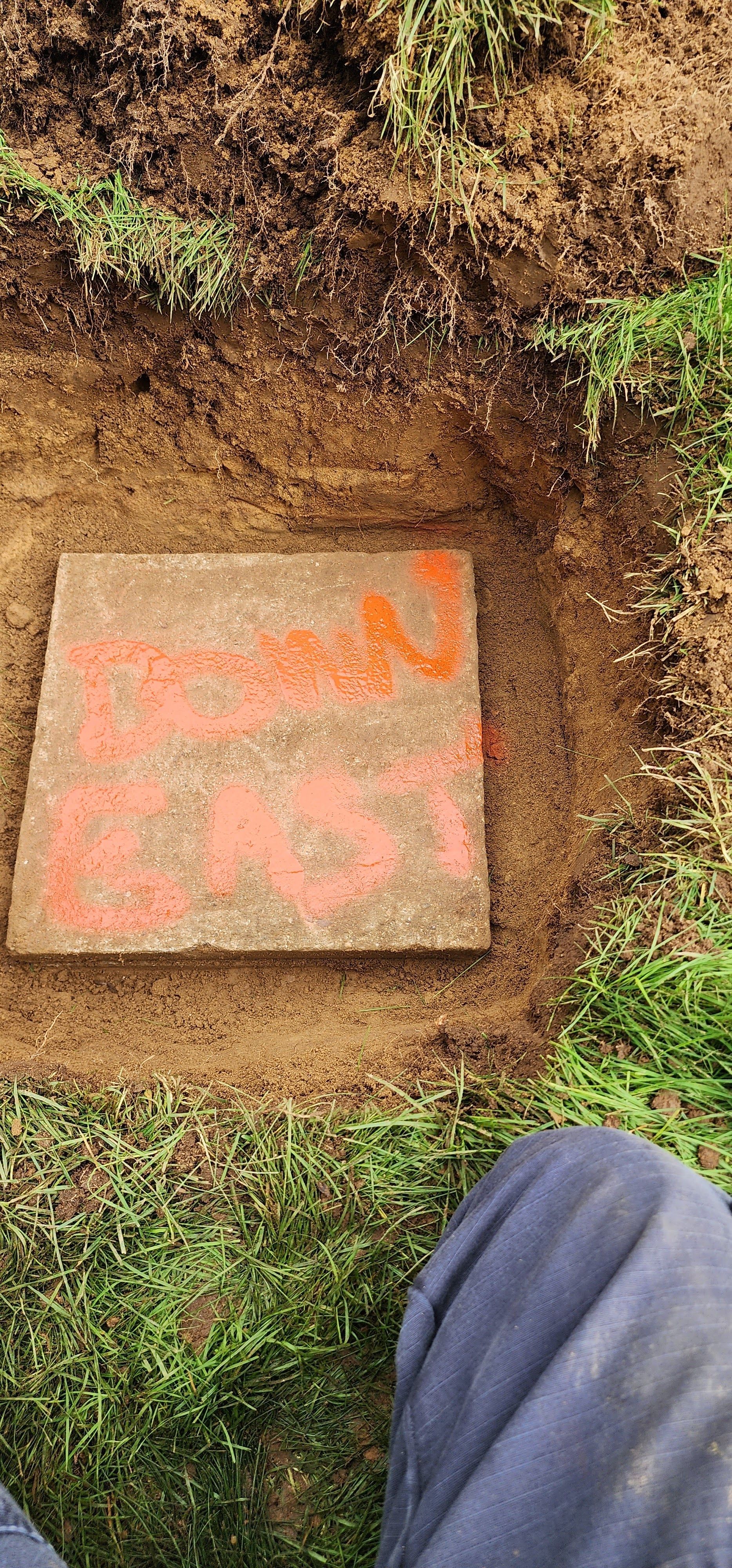 Hand-painted rectangular concrete marker on dirt beside grass, with orange markings and a person’s leg visible.