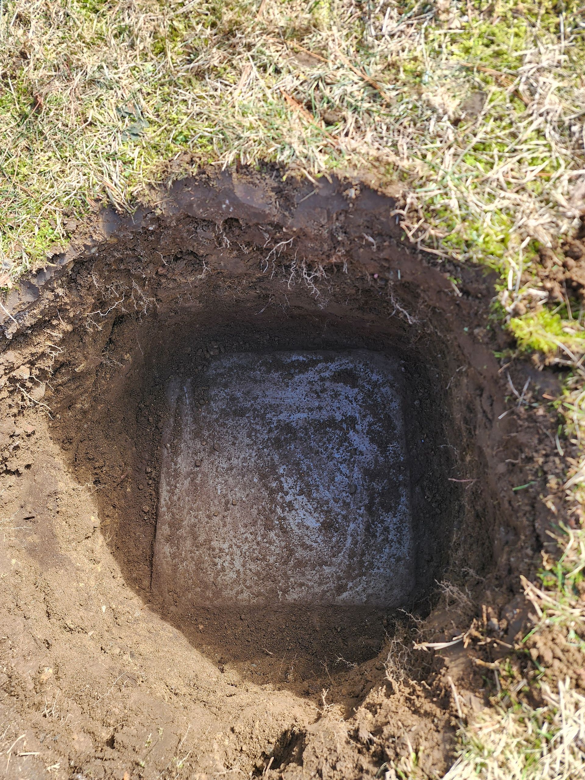 A dug hole in grassy soil, showing a dark, muddy pit with a square-shaped bottom.