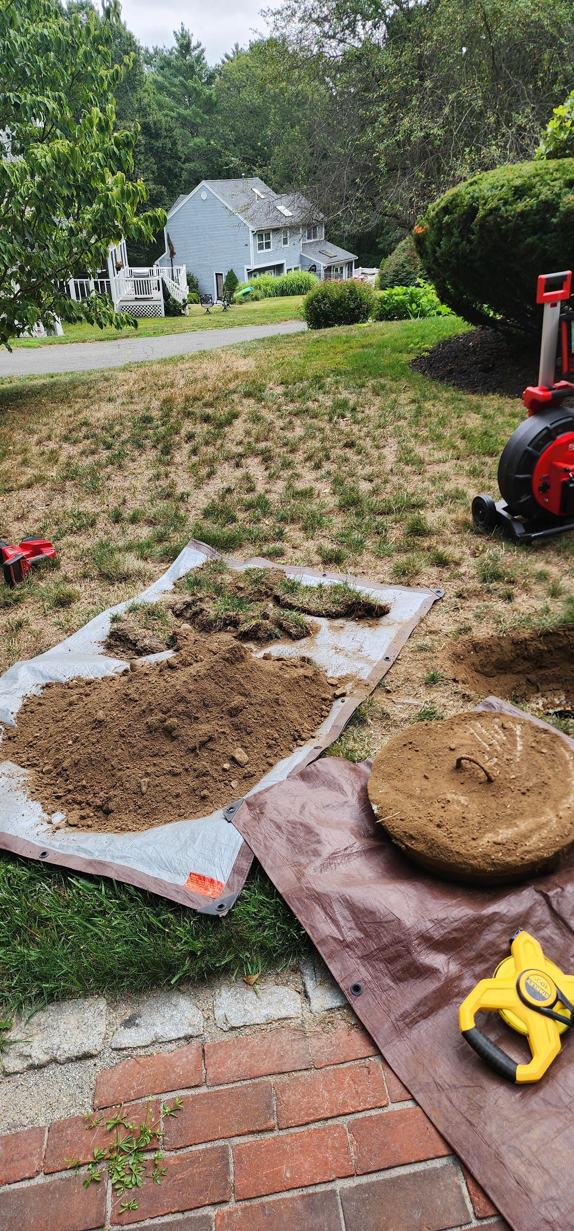 Garden work area with mulch, bricks, a rake, and a red lawn tool on a grassy yard