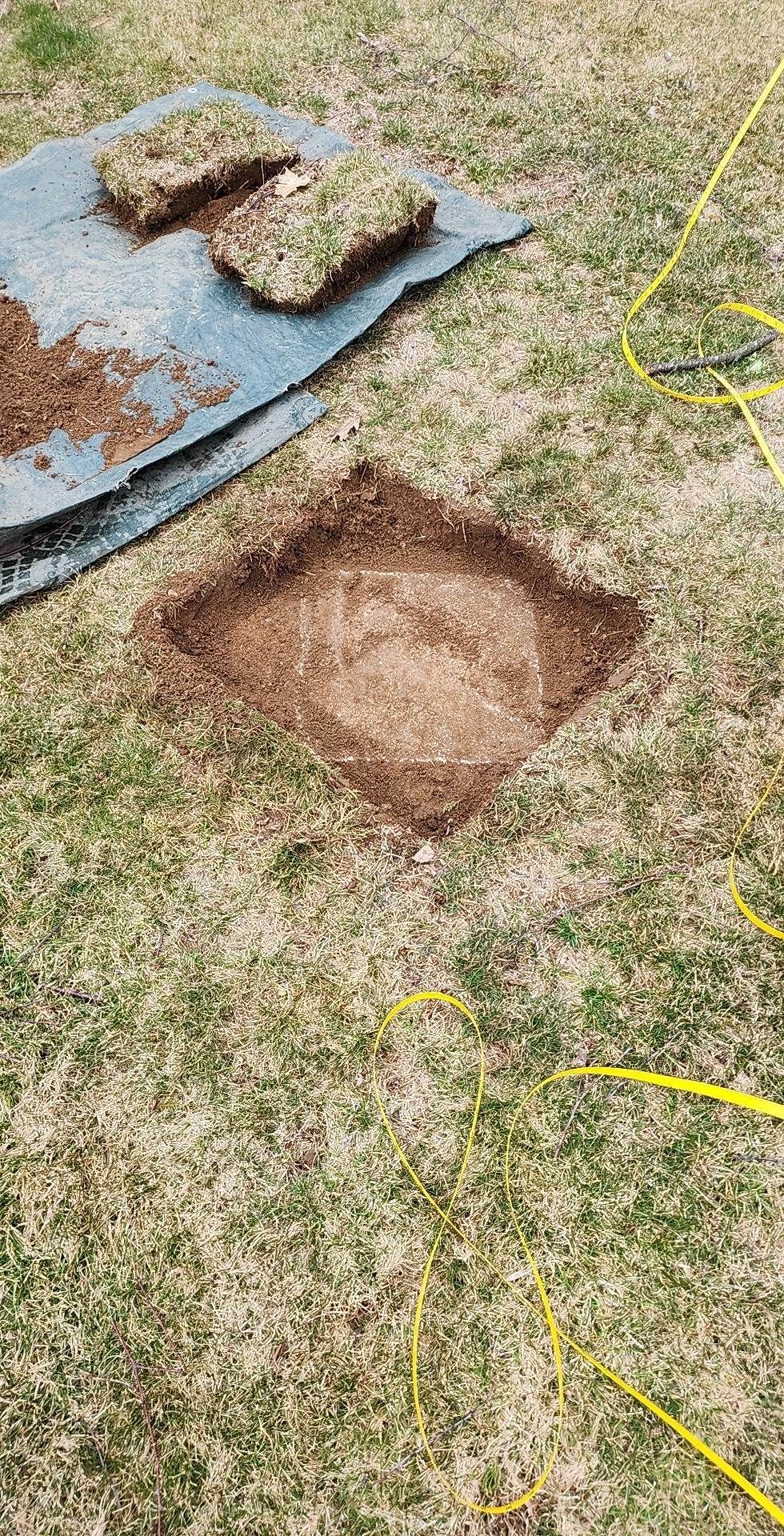 A square hole dug in grass beside a rusty metal sheet and two sod clumps.