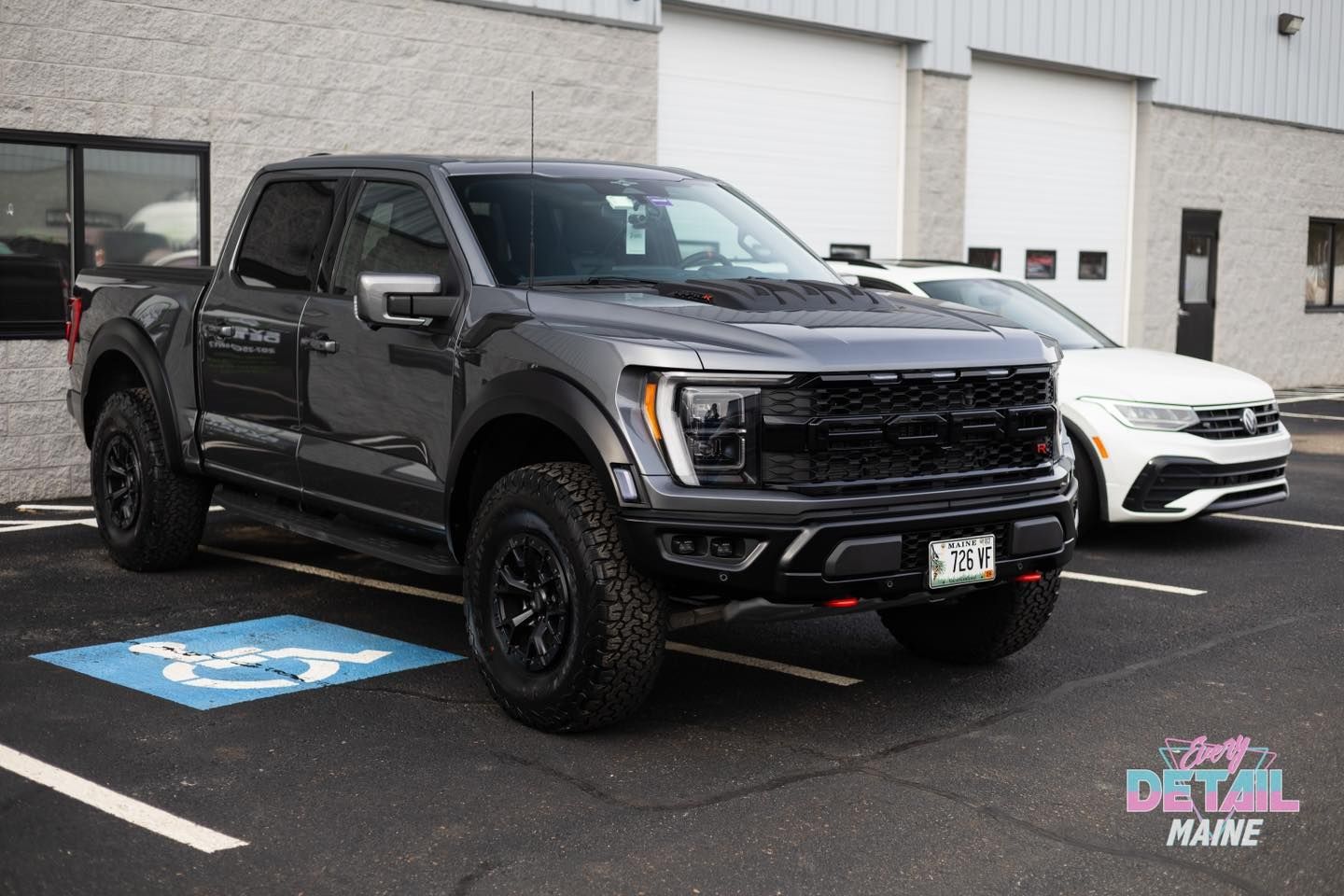 Dark gray Ford Raptor pickup truck parked next to a white car in front of a building; a handicap parking sign is visible.