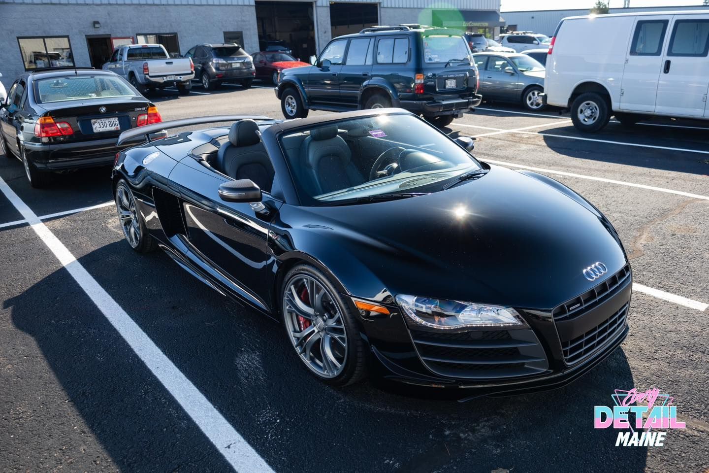 Black Audi R8 Spyder convertible parked in a lot, surrounded by other vehicles.