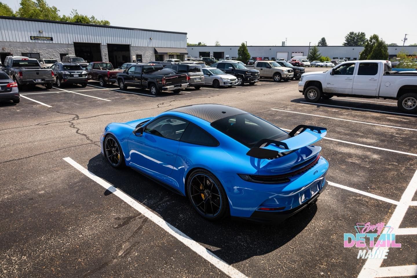 Blue Porsche sports car parked in a lot, black spoiler, sunny day.