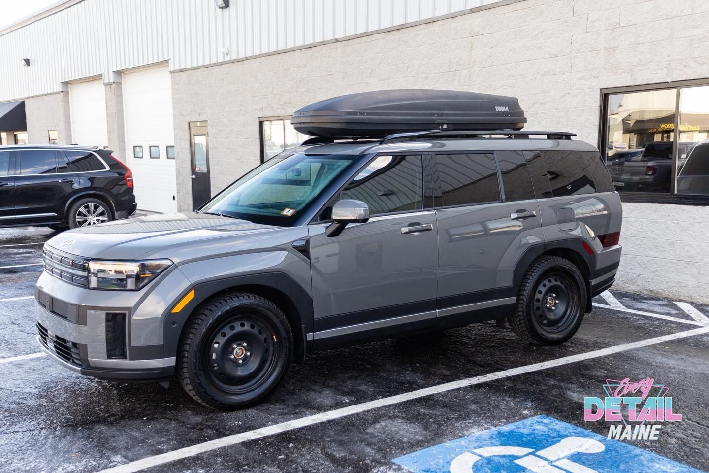 Gray SUV with black roof cargo carrier parked near a building with a handicap parking spot.