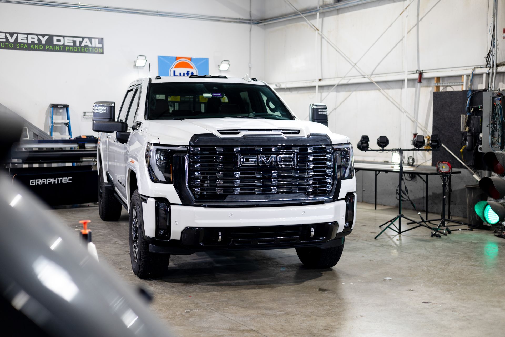 White GMC truck inside a workshop, with black grille and details. "Every Detail" sign visible.