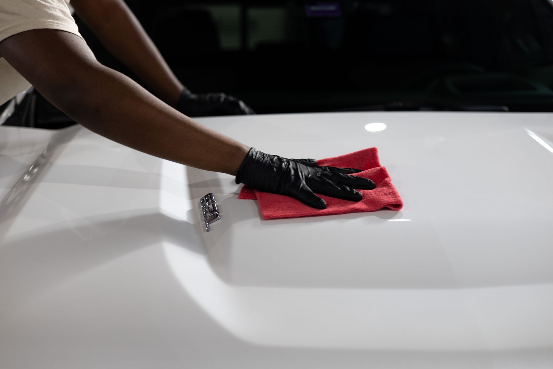 Person wearing black gloves wiping a white car hood with a red cloth.