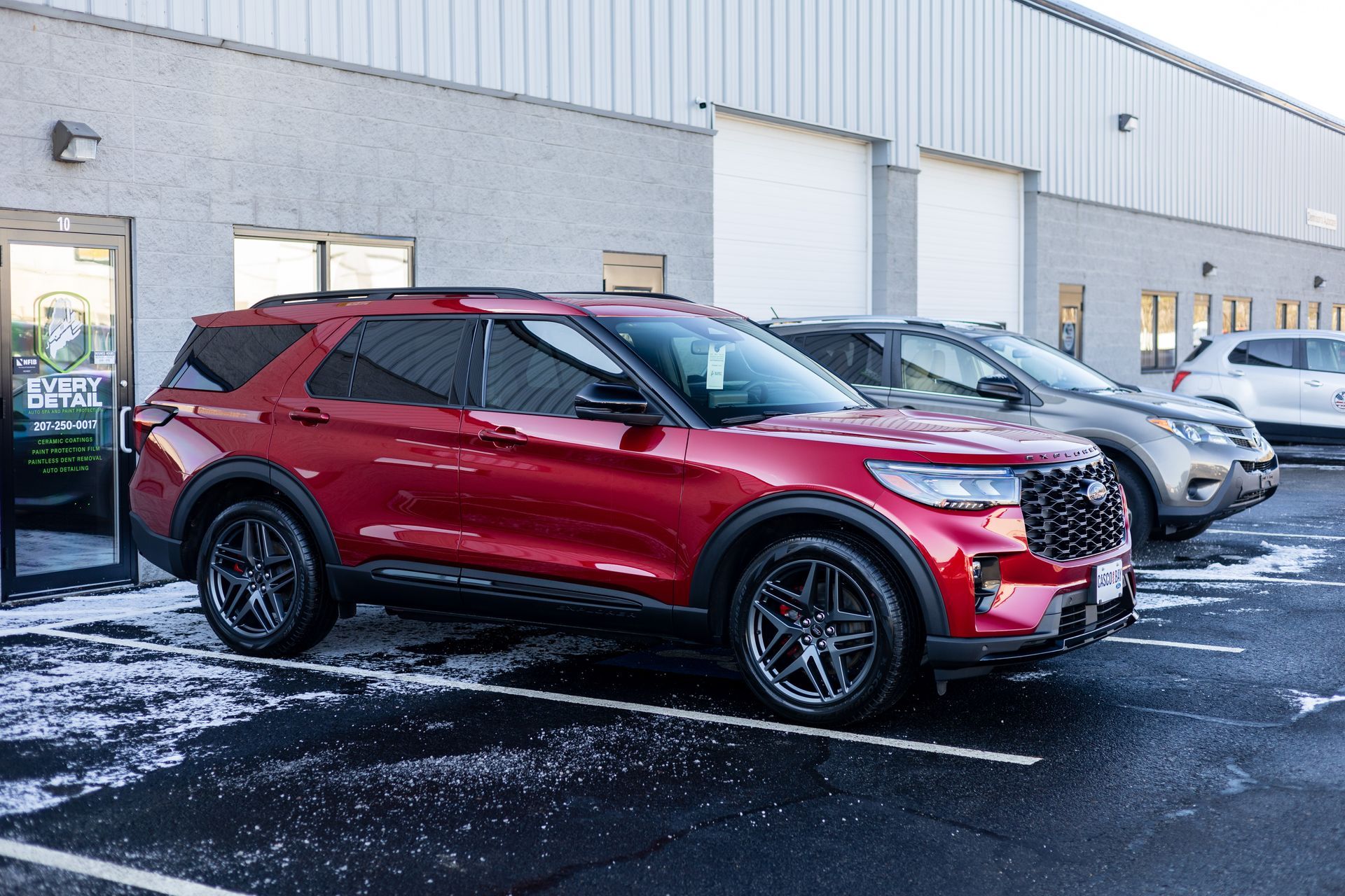 Red Ford Explorer SUV parked in front of a building with other vehicles, gray and white. Snowy ground.