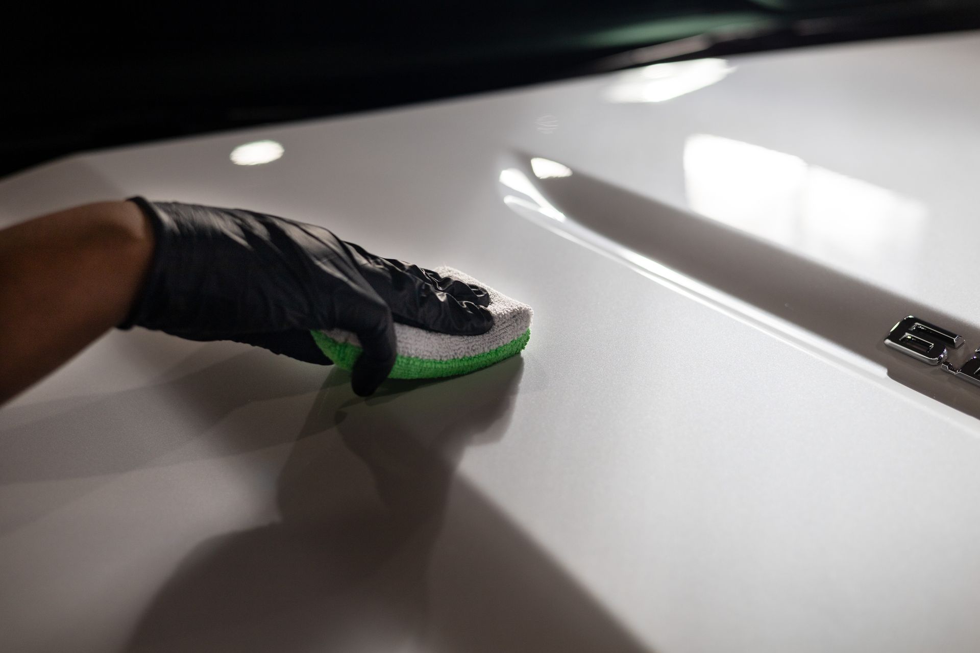 Gloved hand applying a ceramic coating to a white car hood with an applicator sponge.