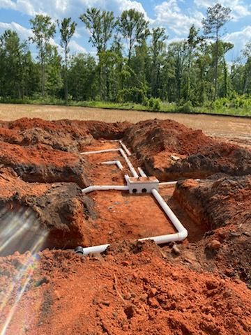 Underground plumbing system installation in red clay soil, with white pipes and a central junction box.
