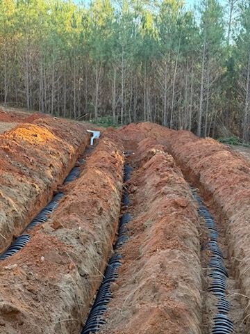 Trenches with black pipes are dug in reddish soil, with trees in the background.