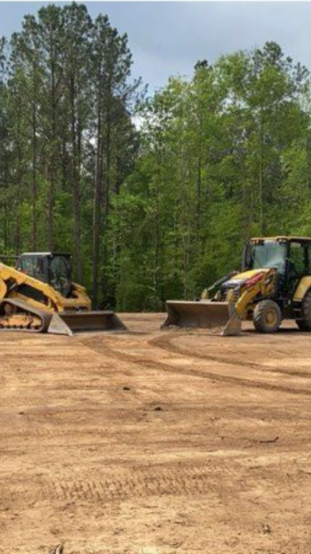 Two yellow construction vehicles on a dirt lot, forest in the background.