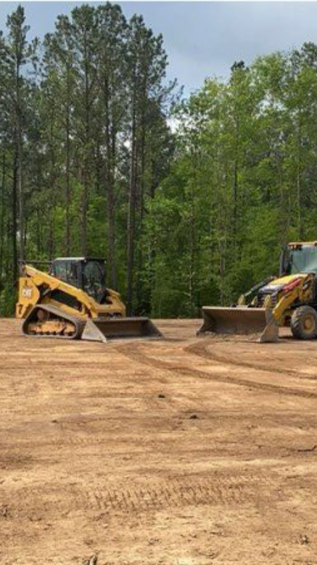 Two yellow construction vehicles on a dirt surface, with a backdrop of trees.