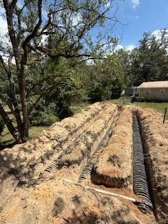 Trenches dug in a yard, containing black drainage pipes, surrounded by dirt and sand, under a blue sky.