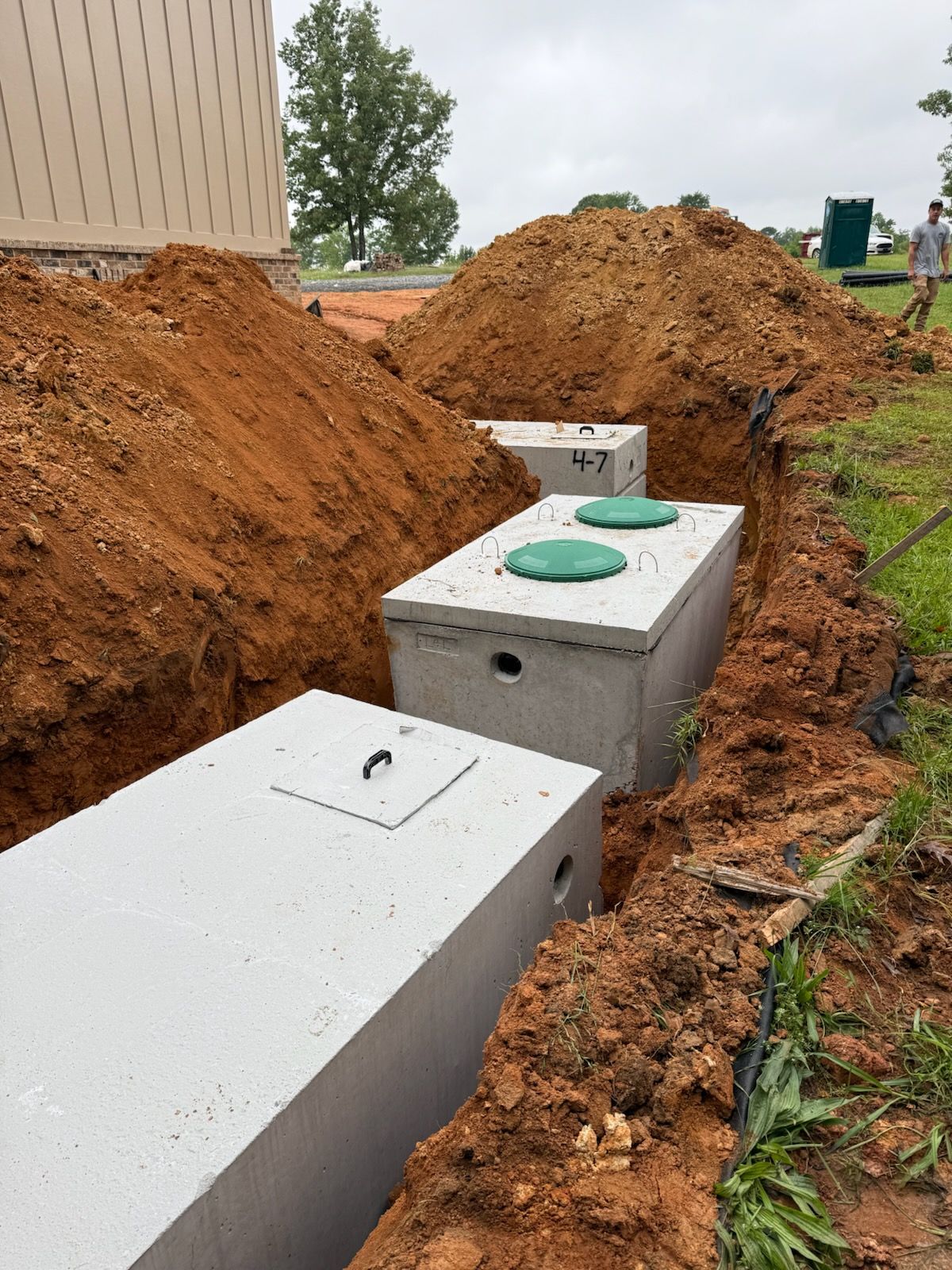 Three concrete septic tanks in a trench, earth piled nearby; grass and a building in the background.