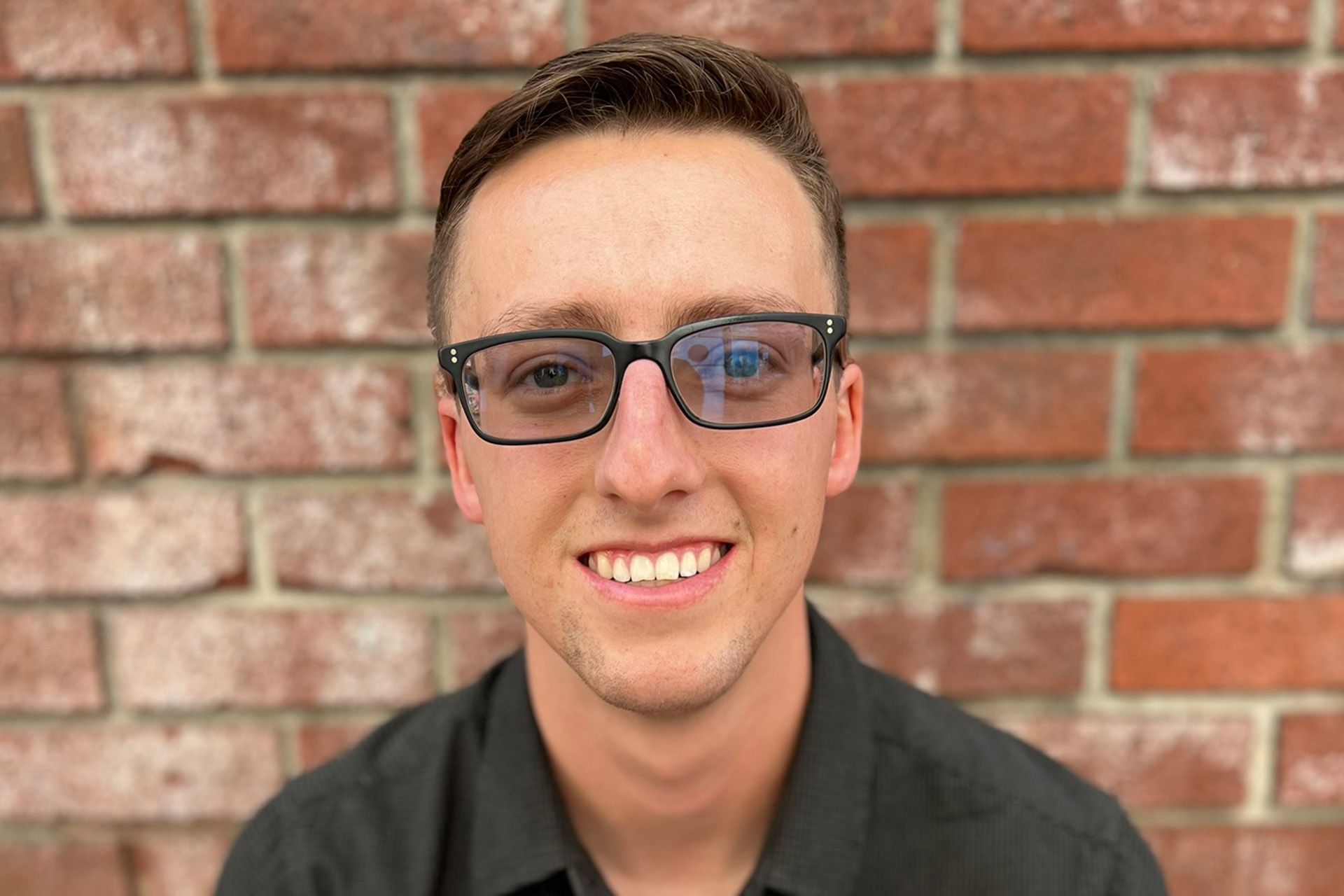 A young man wearing glasses and a black shirt is smiling in front of a brick wall.