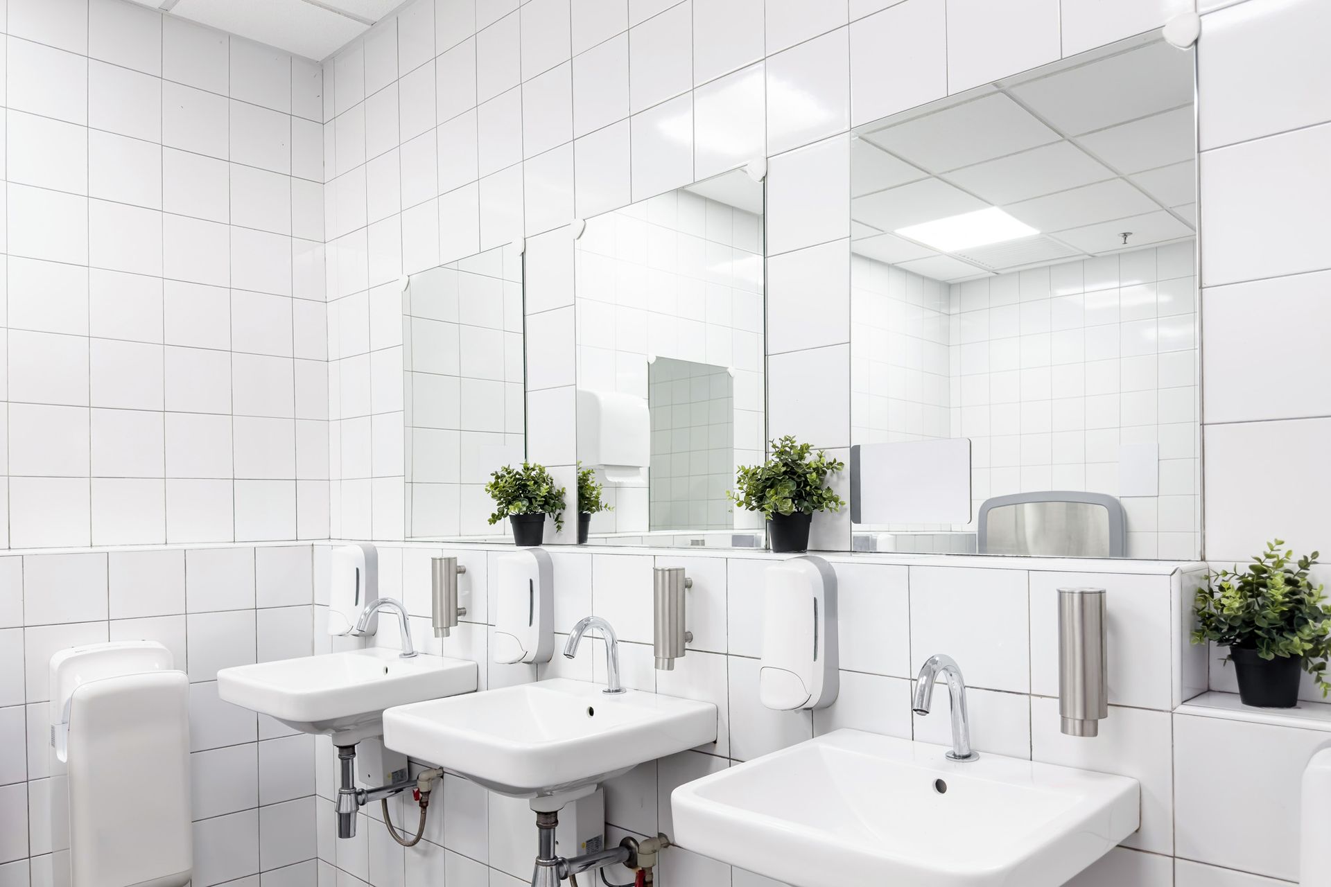 A bathroom with white tiles , sinks , mirrors and plants.