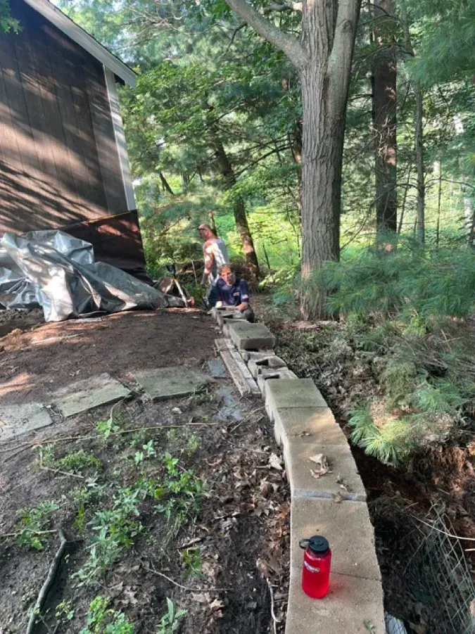 A red water bottle is sitting next to a brick wall in the woods.