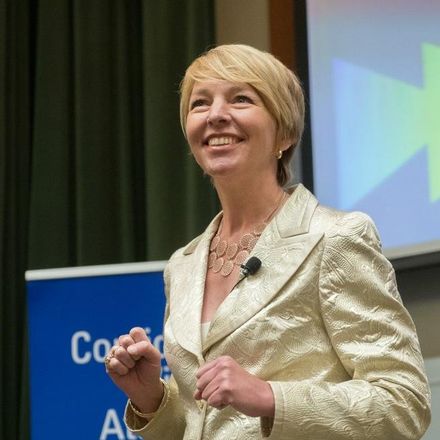 A woman in a gold jacket stands in front of a blue sign that says coa