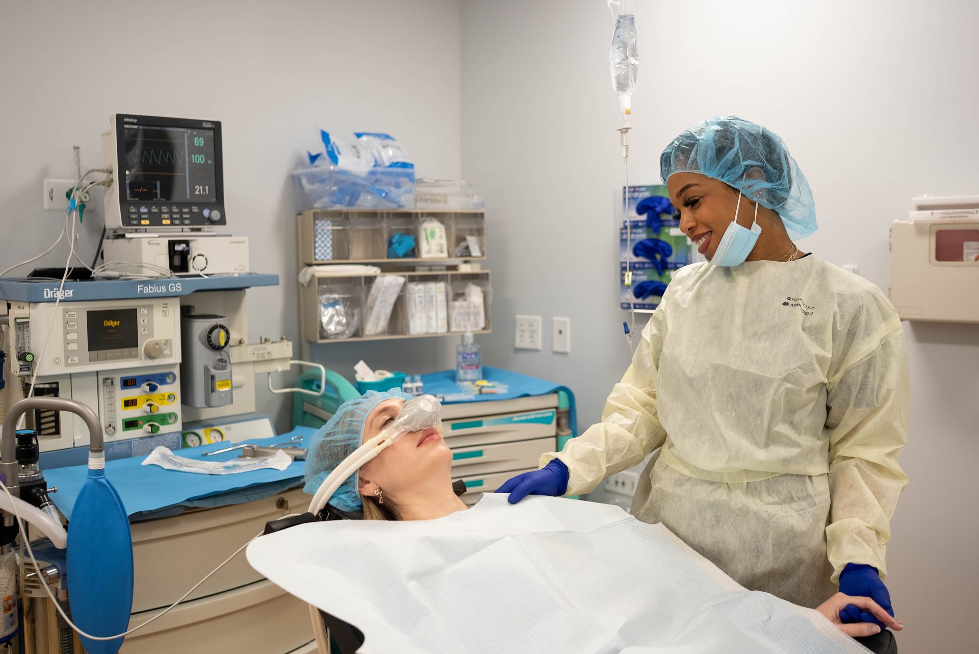 Nurse attends to patient in operating room, patient wears oxygen mask, equipment visible.