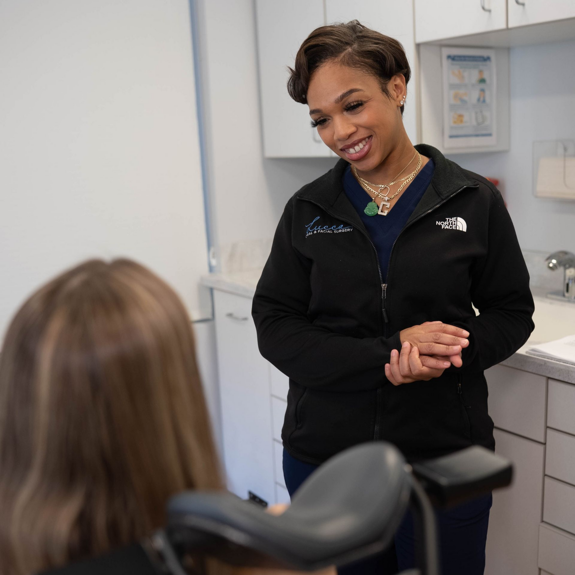 Woman in black jacket smiles, speaking to a person seated in medical chair.