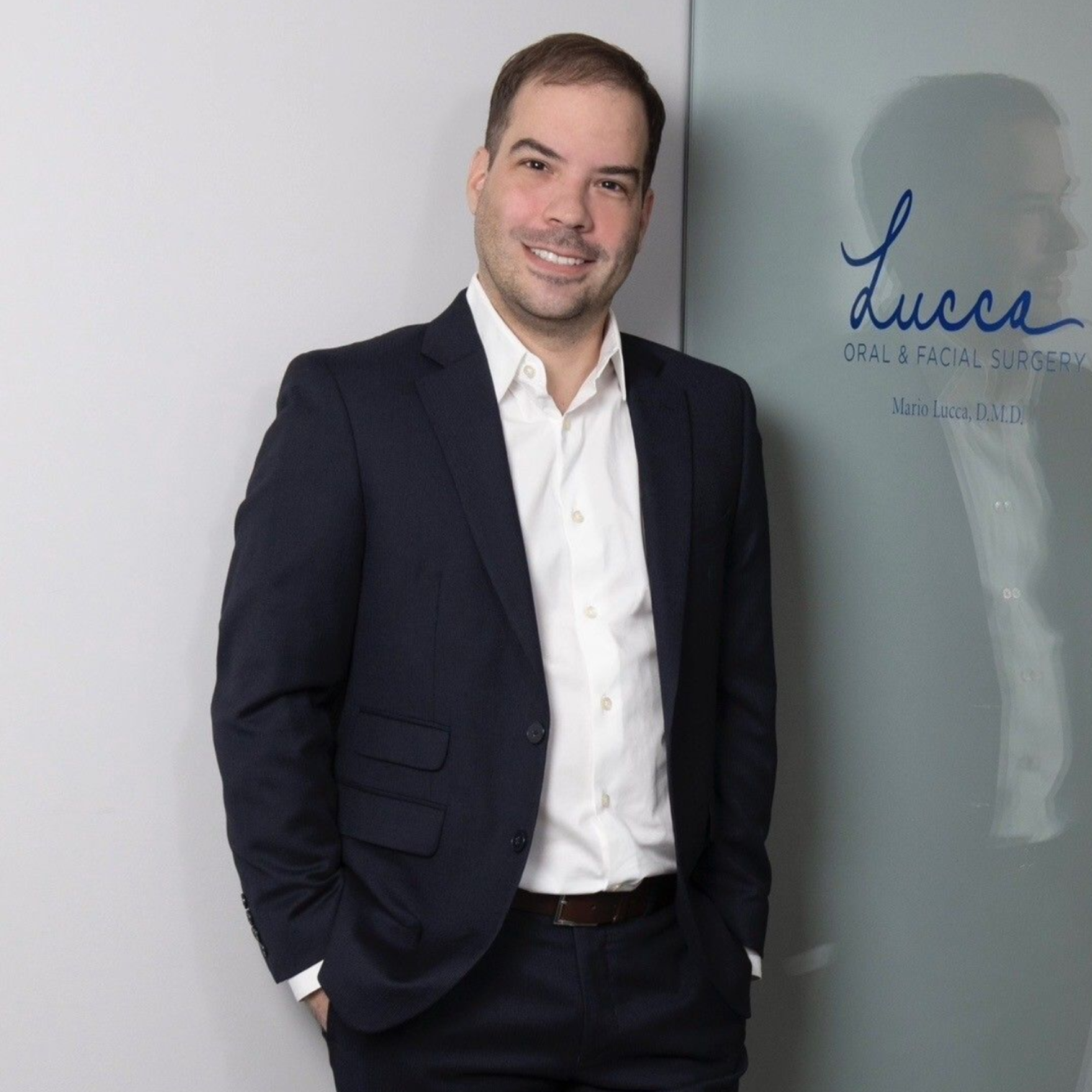 Dr. Mario Lucca wearing a dark suit and white shirt leans against a wall with a Lucca sign, smiling at the camera.