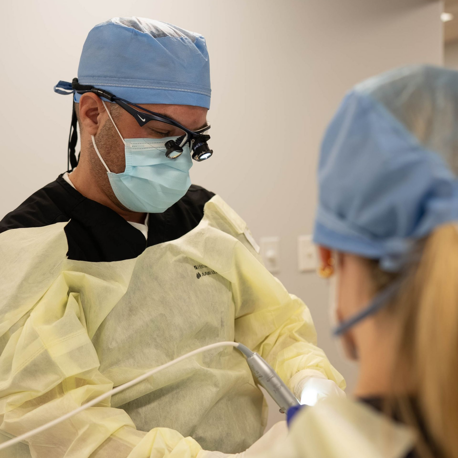 Dr. Lucca in surgical mask and scrubs, working on a patient with an assistant, in a clinic setting.