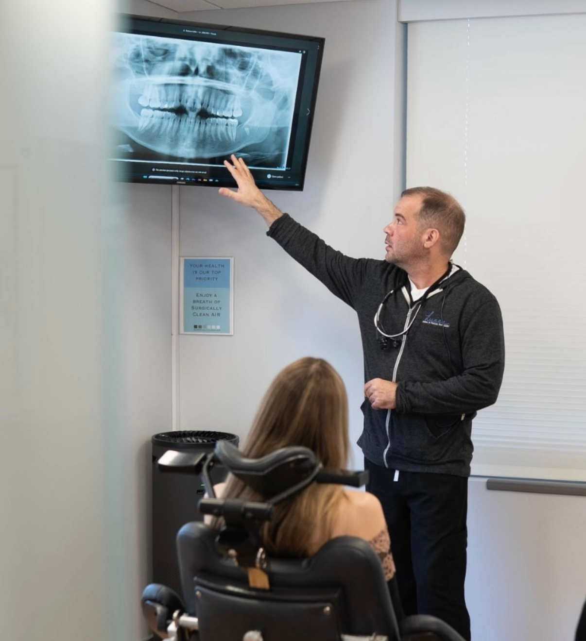 Dr. Lucca in gray hoodie smiling, speaking to patient in dental chair, window behind them with an x-ray.