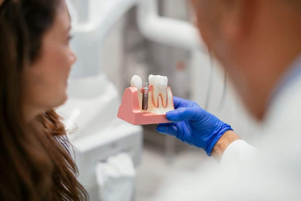 A dentist wearing blue gloves holds a model of a dental implant and teeth to explain the procedure to a patient.