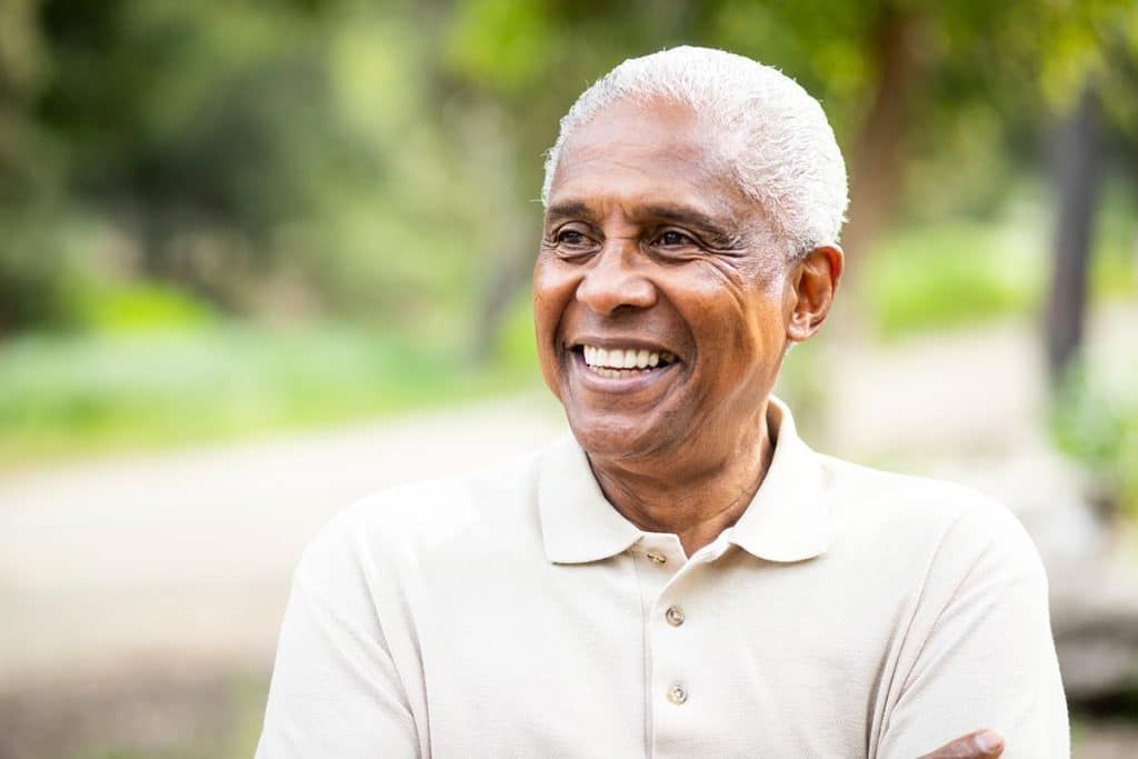 A smiling person with grey hair wears a beige polo shirt outdoors, looking toward the side with arms crossed.