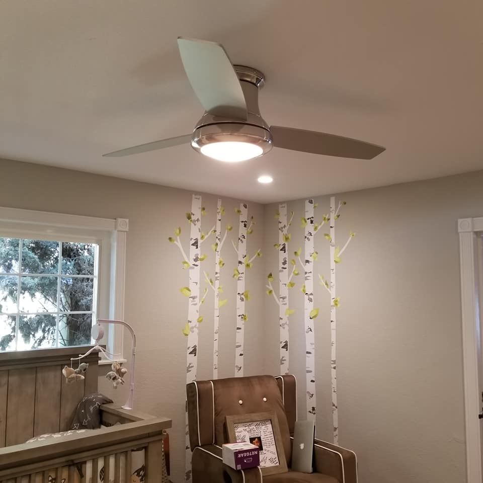 A room with a ceiling fan , chair , crib and window.