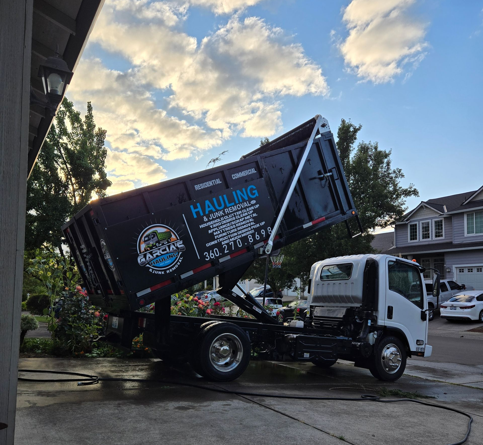 A black pickup truck with a trailer dumping debris on a driveway in front of a house.