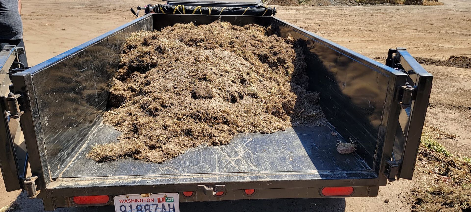 A black trailer filled with brown mulch is on a sunny day. A license plate is visible at the bottom.