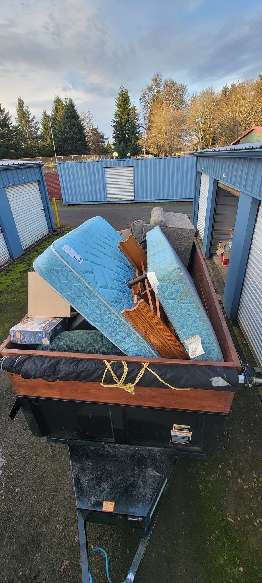 A trailer loaded with trash, including a ripped blue mattress, sits in front of storage units.