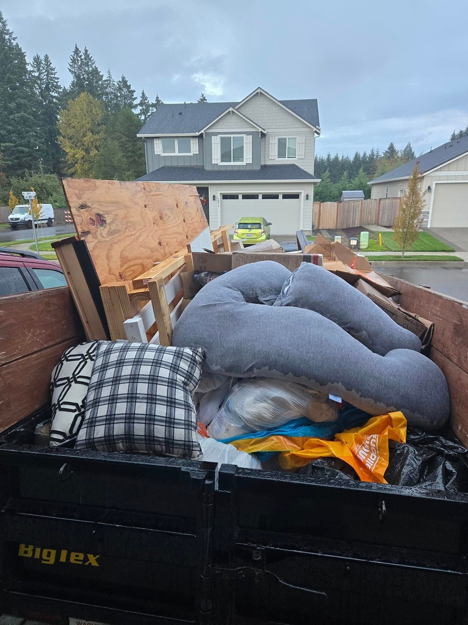 A dumpster overflowing with household items, including a gray pillow, in front of a two-story house.