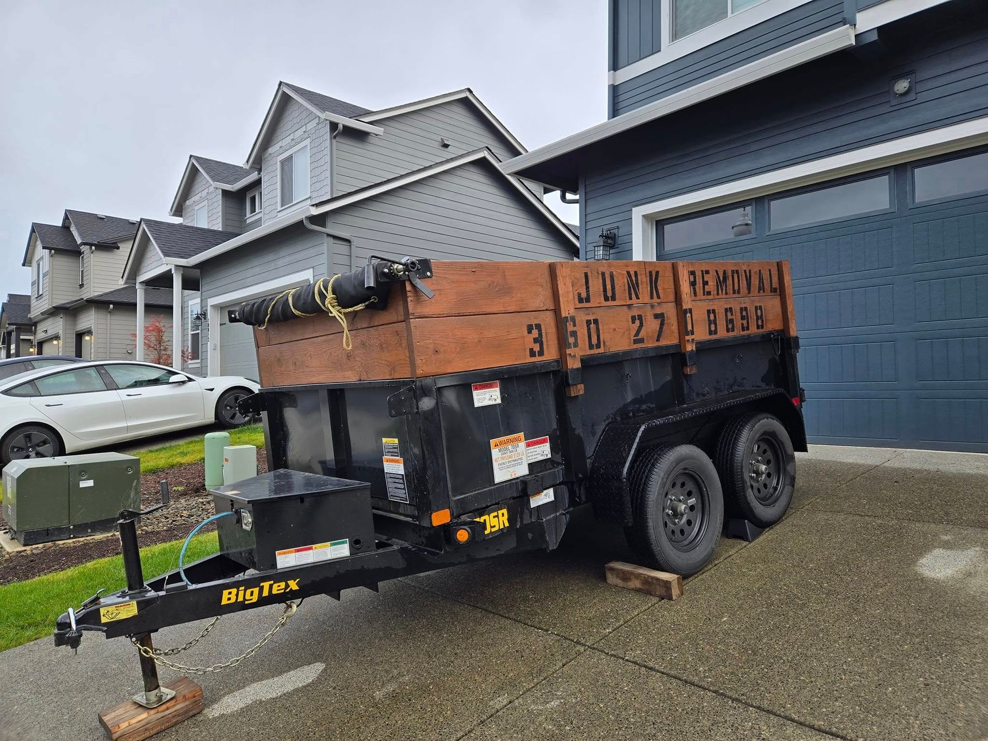 Black Rig Flex trailer filled with brown wood, parked in front of a house. A white car is next to the trailer.