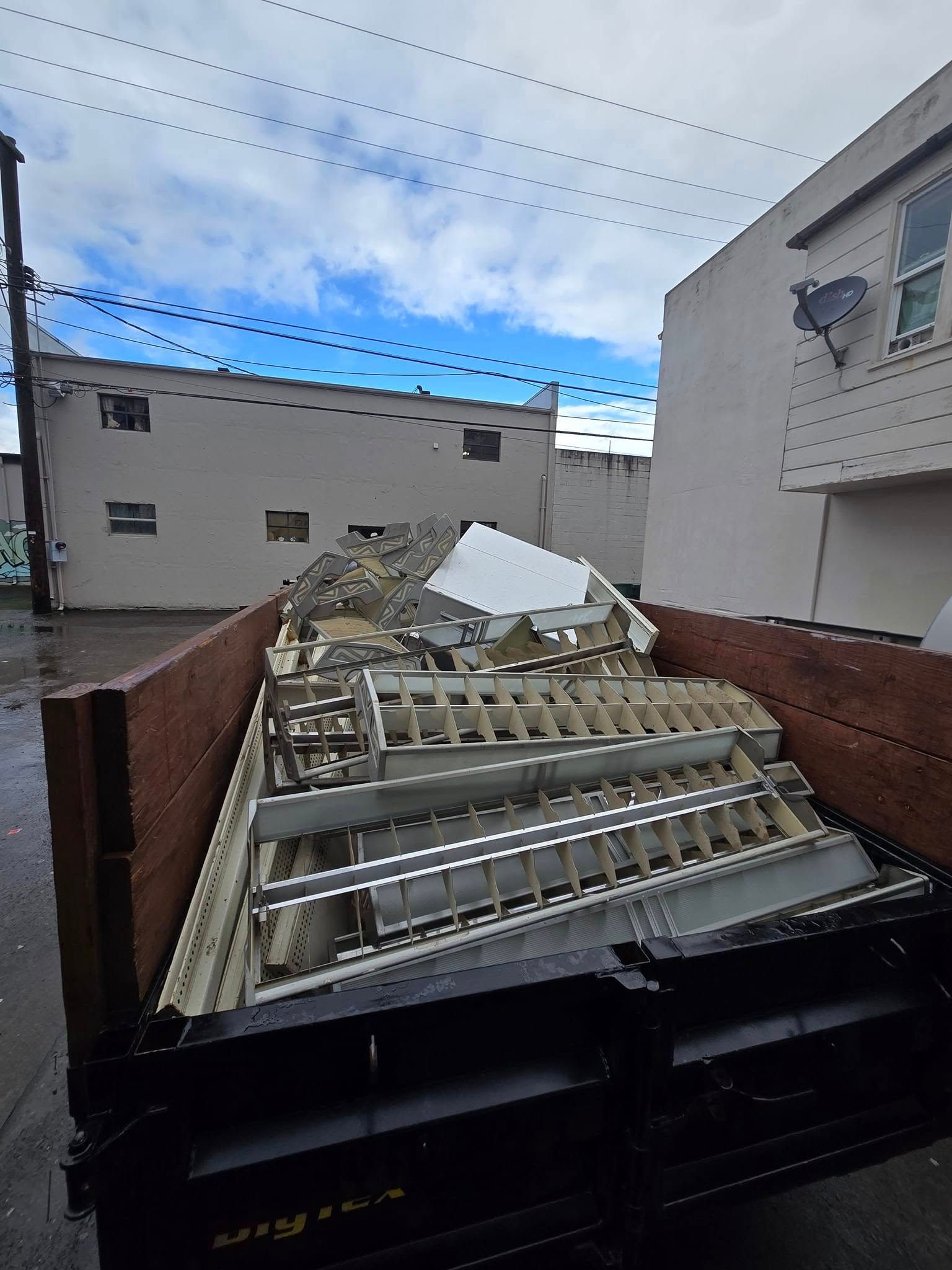 A loaded dumpster filled with metal debris in an alleyway between two buildings, cloudy sky overhead.