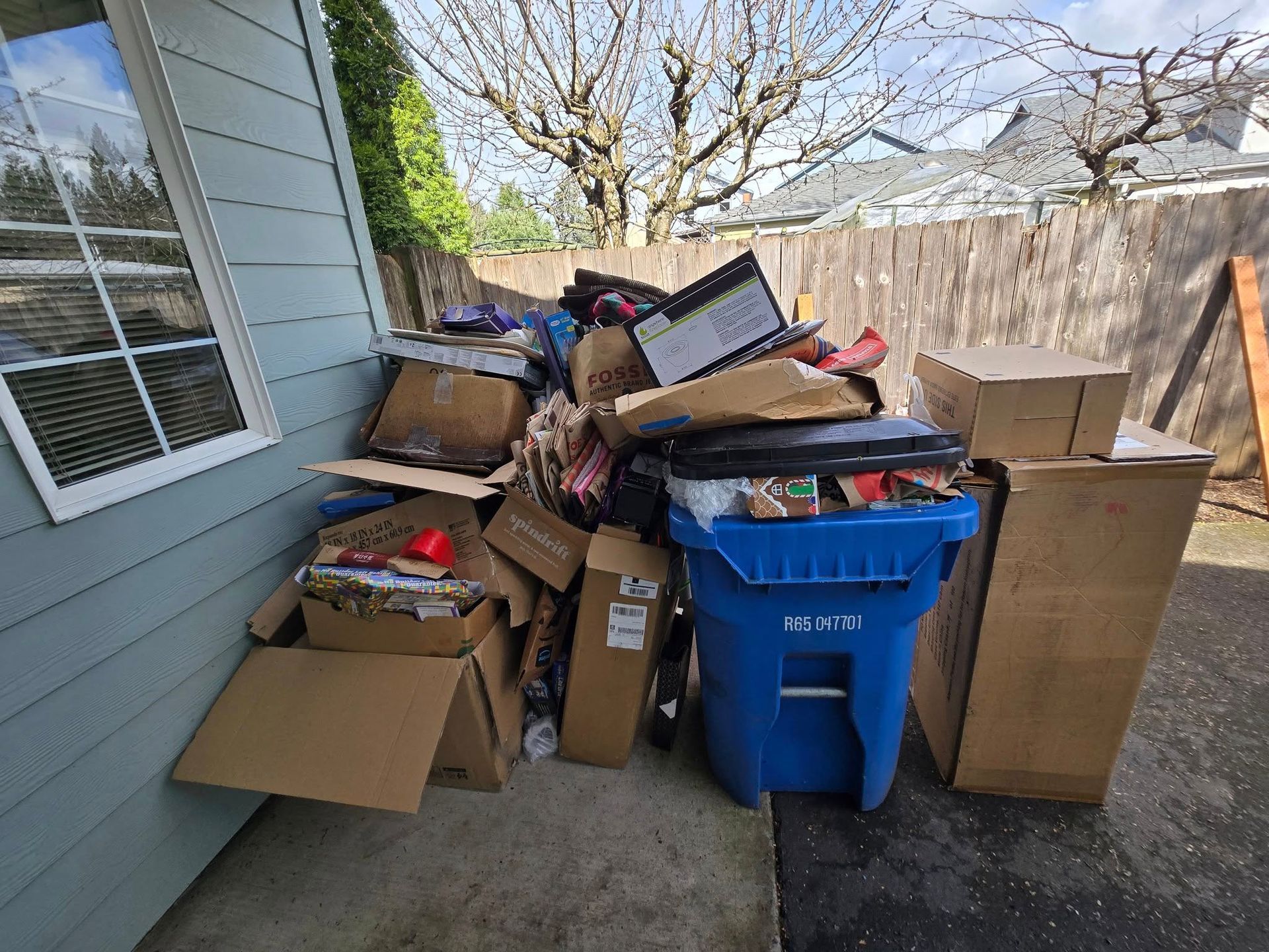 A pile of cardboard boxes, trash, and overflowing blue recycle bin next to a house and fence.