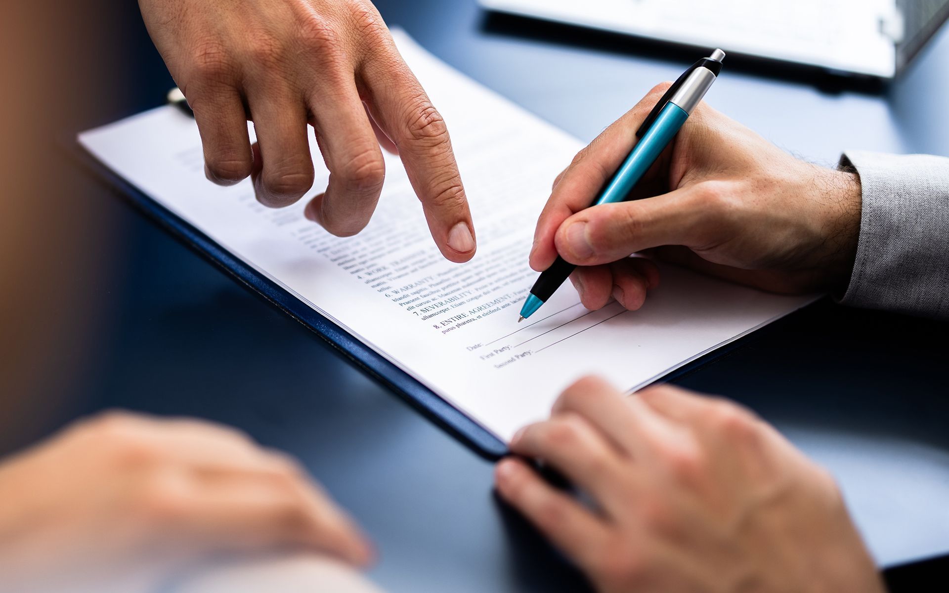 Two hands signing a document on a clipboard; another hand points to the signature line.