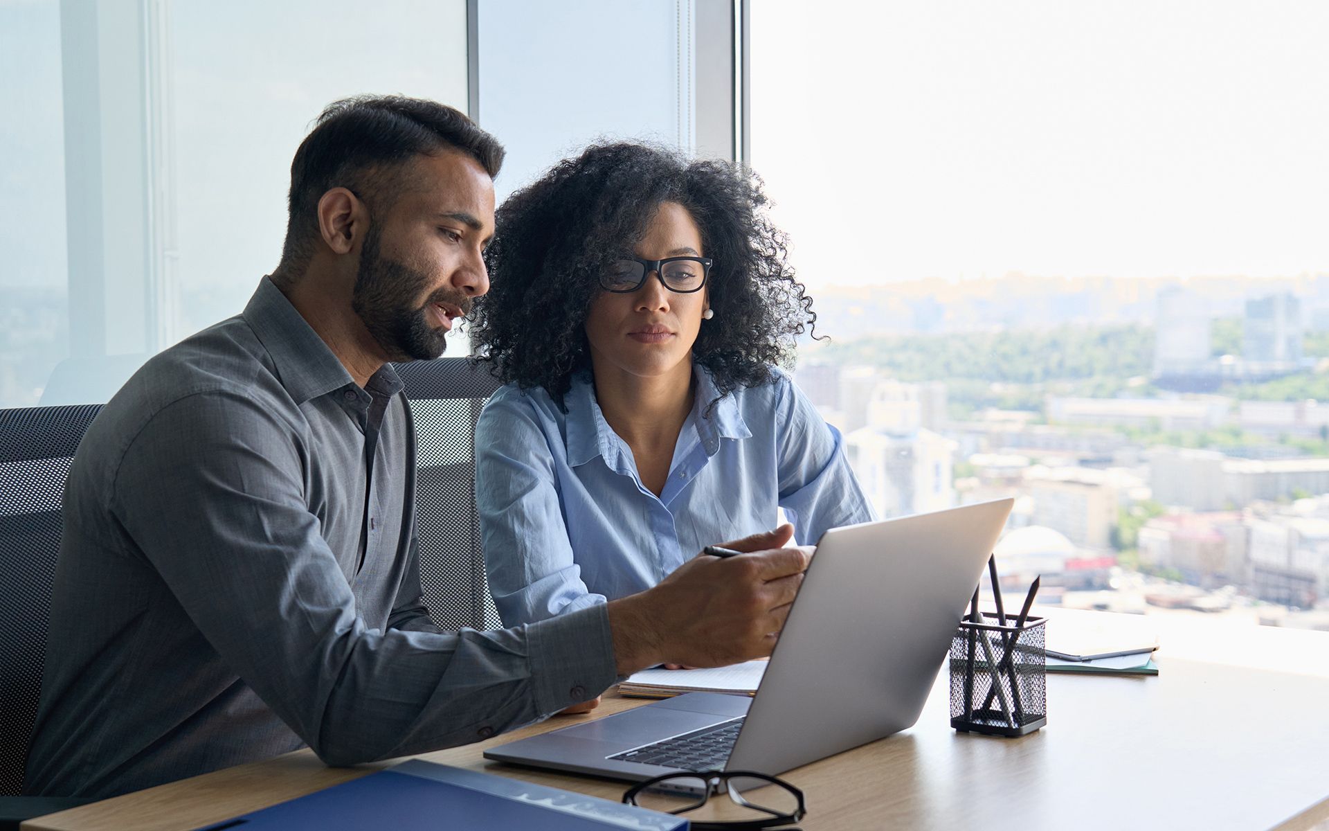 Man and woman reviewing laptop together at a desk near a window in an office.