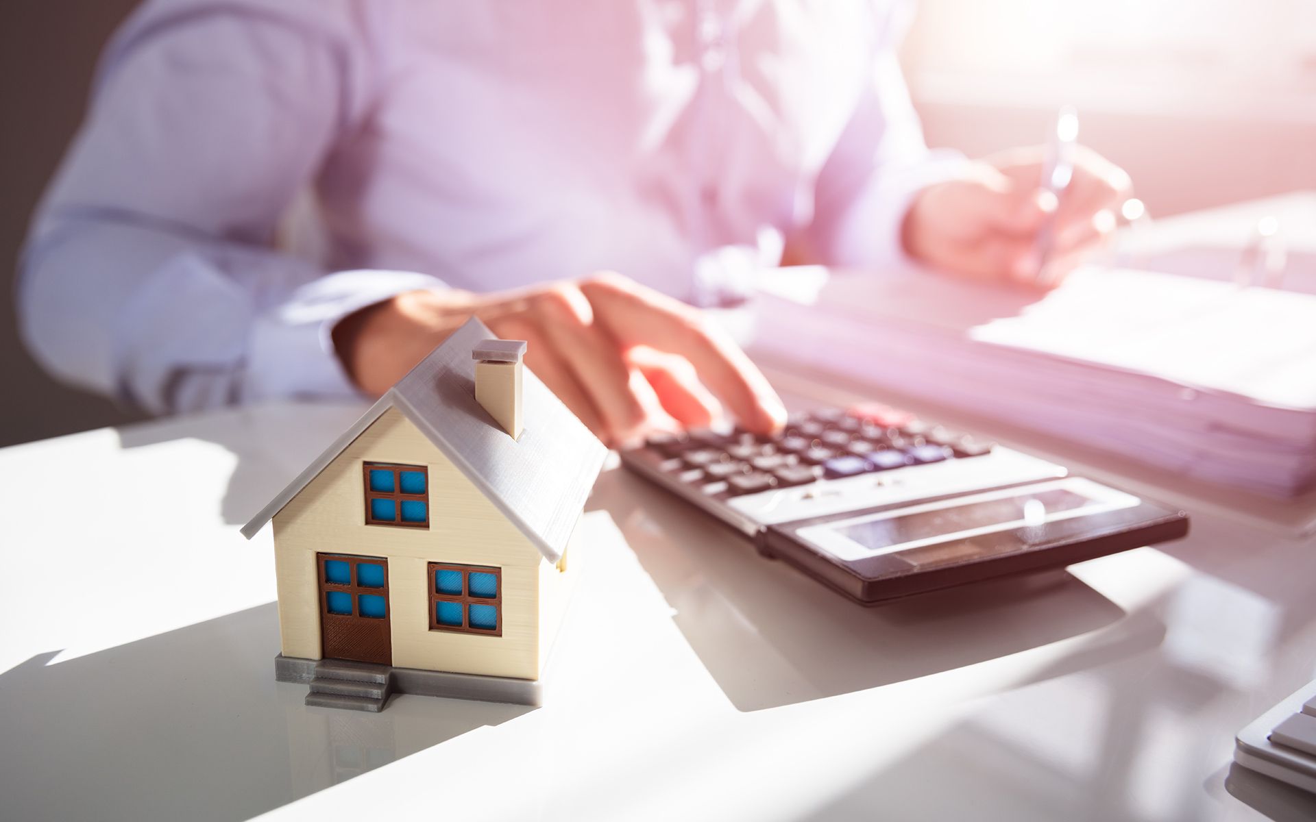 Person calculating with a calculator next to a miniature house model, documents, and sunlight.