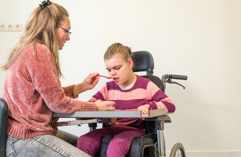 A Woman is Feeding a Young Girl in a Wheelchair With a Spoon — Specialist Support Solutions in Rockhampton City, QLD