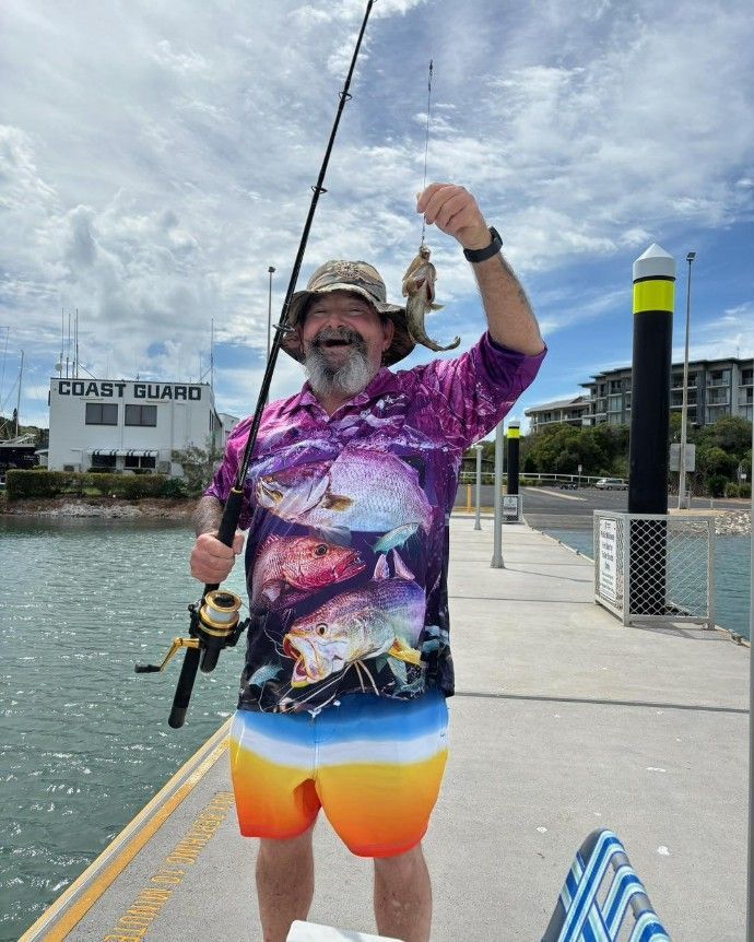 A Man is Standing on a Dock Holding a Fishing Rod and a Fish — Specialist Support Solutions in Rockhampton City, QLD